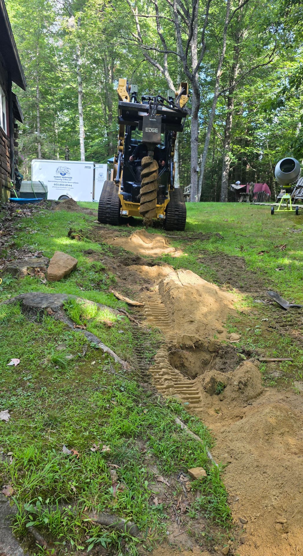 Skid steer auger boring a utility trench beside a log cabin